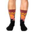 Front view of legs wearing black crew socks with a prominent warm-toned sunset design on the shins.