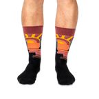 Front view of legs wearing black crew socks with a prominent warm-toned sunset design on the shins.