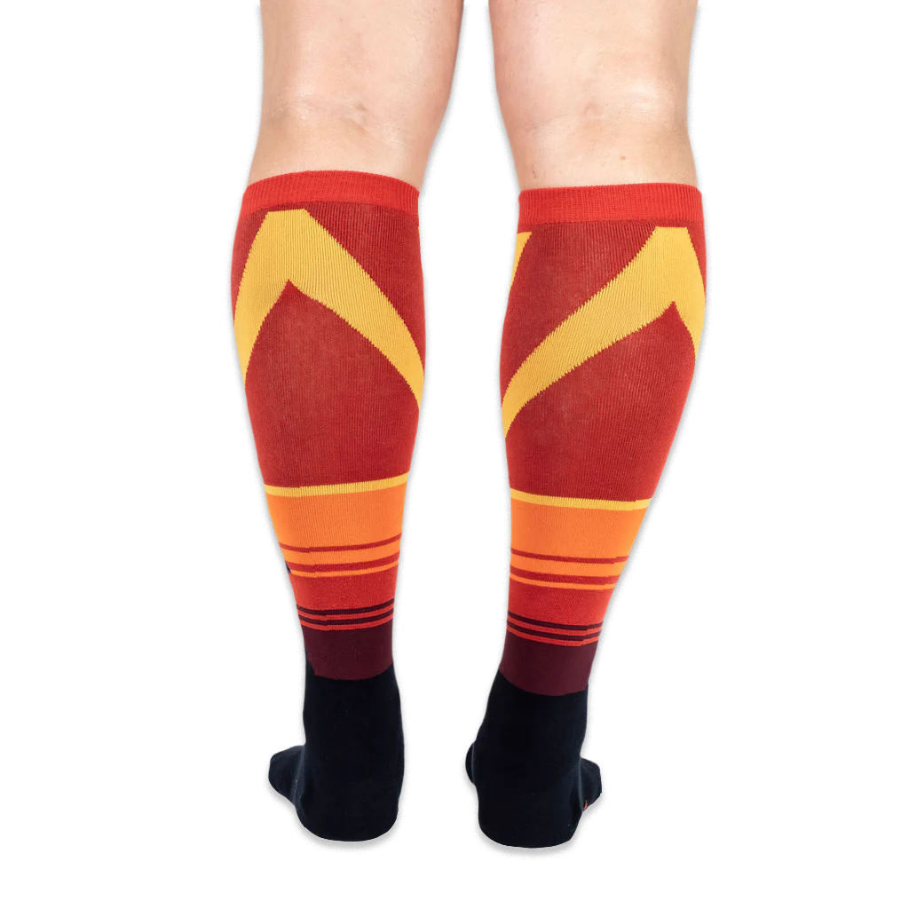 Back view of the knee-high socks being worn, displaying a bold yellow chevron pattern over horizontal orange stripes.