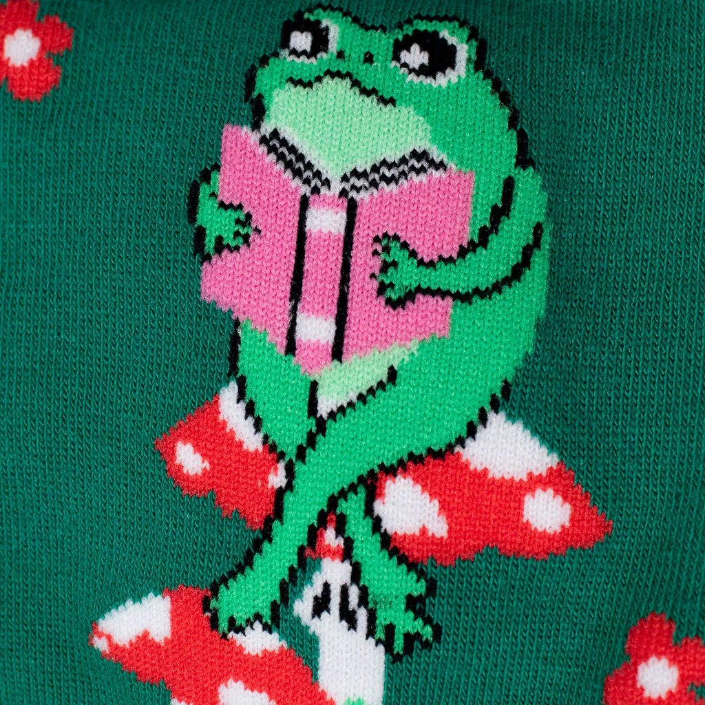 Close-up knitted detail of a green frog sitting on a red and white spotted mushroom while reading a bright pink book.