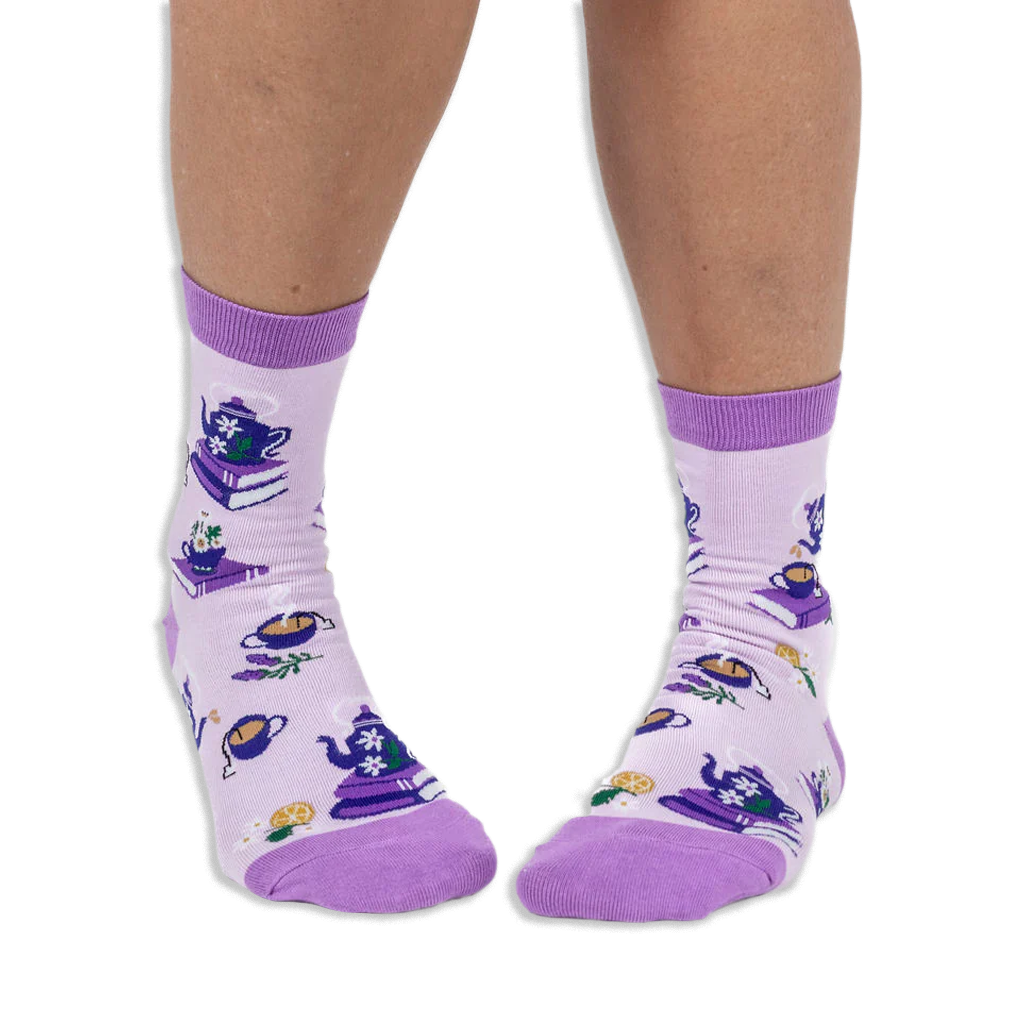 Front view of a person wearing light purple crew socks showing a pattern of teapots on books and steaming cups of tea.