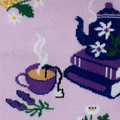 Close-up knit detail of a purple steaming teacup with a tea bag tag, a sprig of lavender, and a blue teapot on purple books.