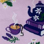 Close-up knit detail of a purple steaming teacup with a tea bag tag, a sprig of lavender, and a blue teapot on purple books.