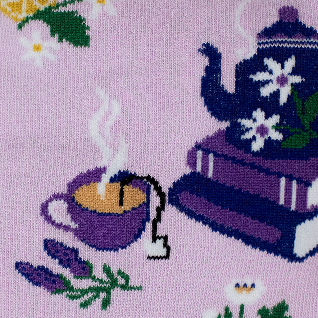 Close-up knit detail of a purple steaming teacup with a tea bag tag, a sprig of lavender, and a blue teapot on purple books.