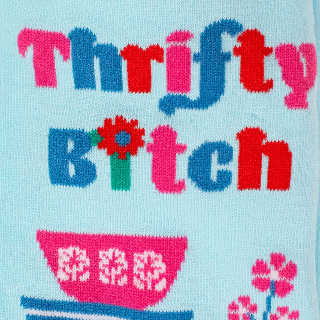 Close-up detail of knitted Thrifty B*tch text in pink and blue with red accents, plus a pink patterned mixing bowl.