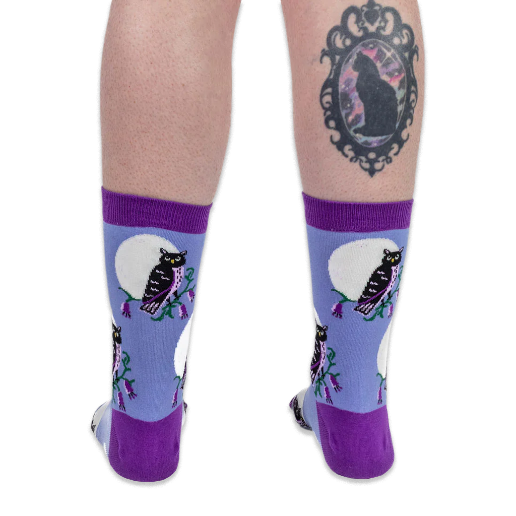 Back view of legs wearing blue owl socks. The right calf features a black cat cameo tattoo on a solid black background.