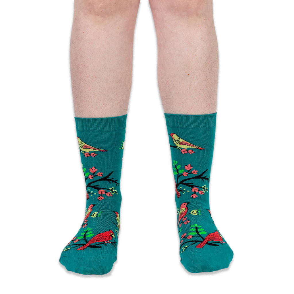 Front view of a person's lower legs wearing teal crew socks with a pattern of birds, black branches, and pink flowers.