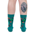 Back view of legs wearing teal bird socks. The person has a large framed black cat tattoo on their right calf.