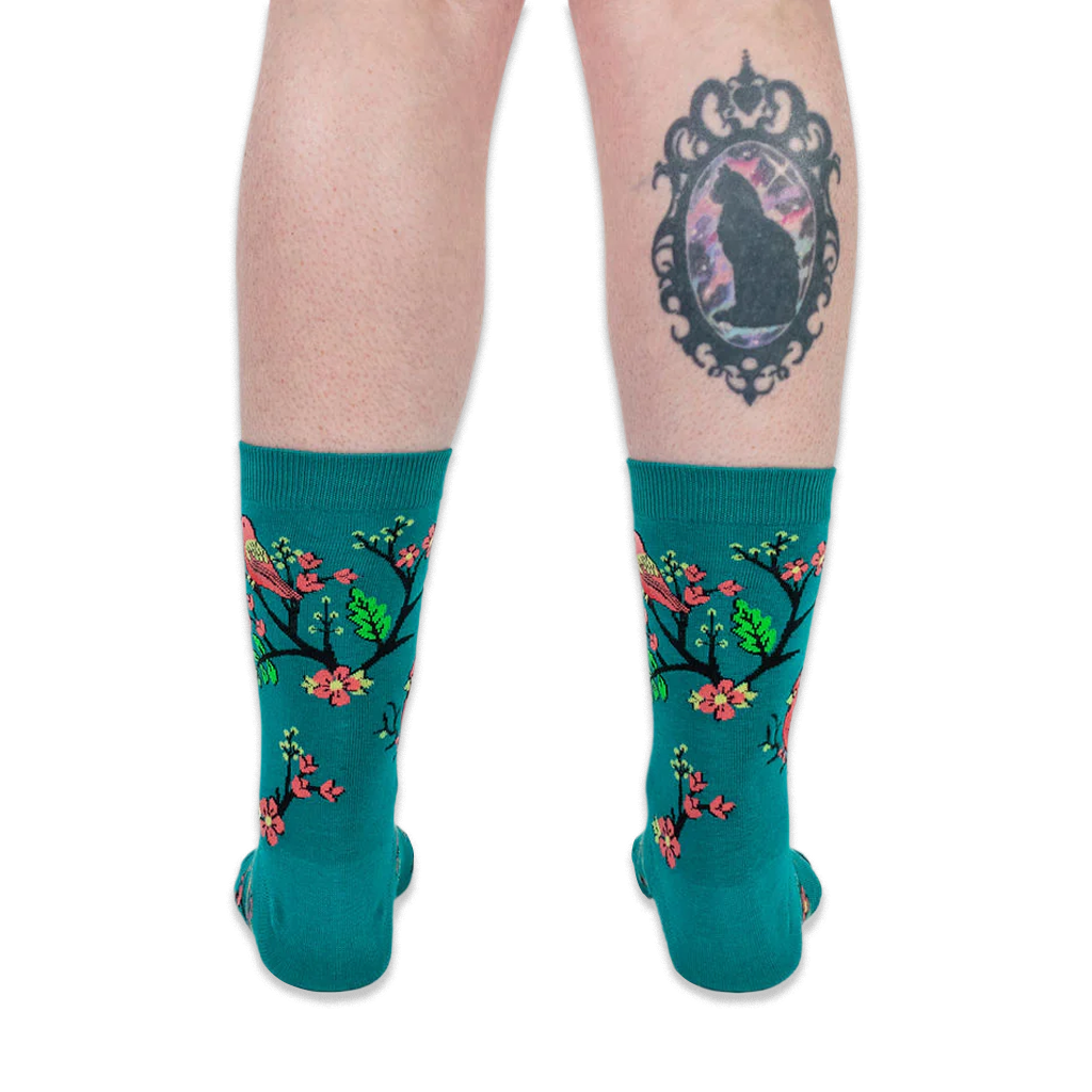 Back view of legs wearing teal bird socks. The person has a large framed black cat tattoo on their right calf.