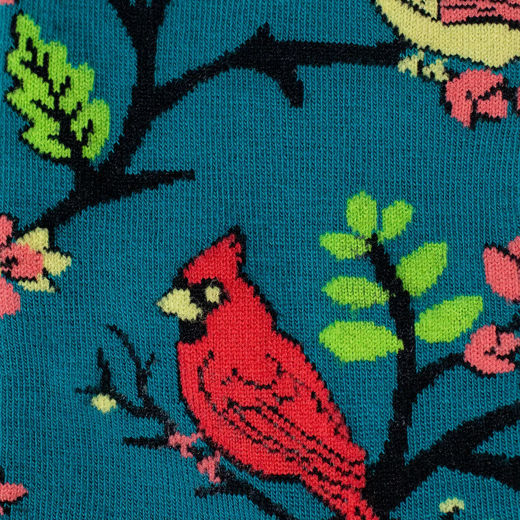 Close-up knit detail of a red cardinal on a black branch with bright green leaves against a textured teal background.