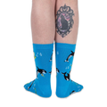 Back view of a person wearing bright blue musical orca socks, showing a black cat tattoo on the back of their right calf.