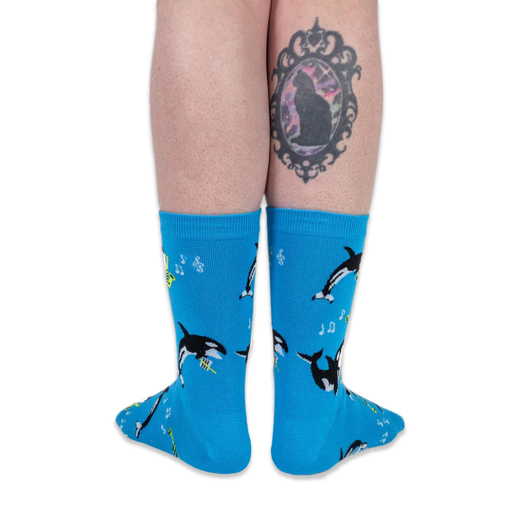 Back view of a person wearing bright blue musical orca socks, showing a black cat tattoo on the back of their right calf.