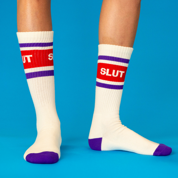 [lf] Person's legs wearing cream crew socks with 'SLUT' graphic, blue-purple stripes, toe, and heel on a blue background.
