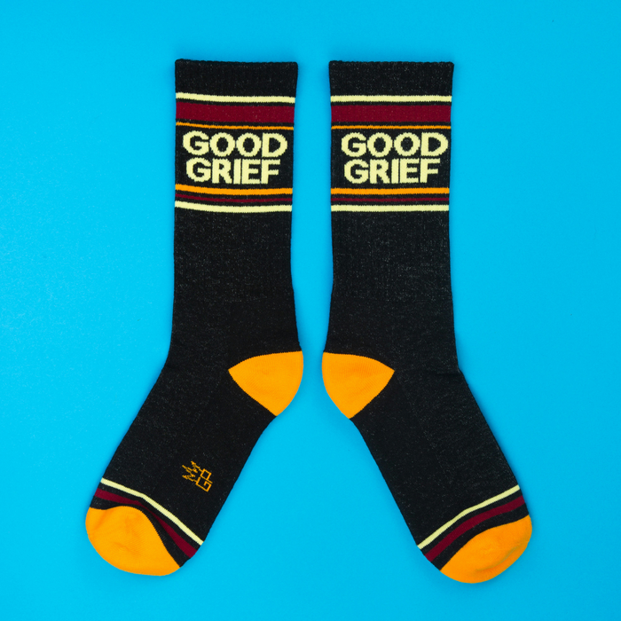 Two black 'Good Grief' crew socks with colorful stripes and orange accents, laid flat on a blue surface.