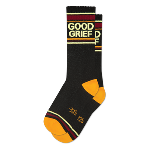 A folded pair of black crew socks featuring 'Good Grief' text, orange toe and heel, and burgundy, gold, and cream stripes.