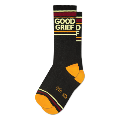 A folded pair of black crew socks featuring 'Good Grief' text, orange toe and heel, and burgundy, gold, and cream stripes.