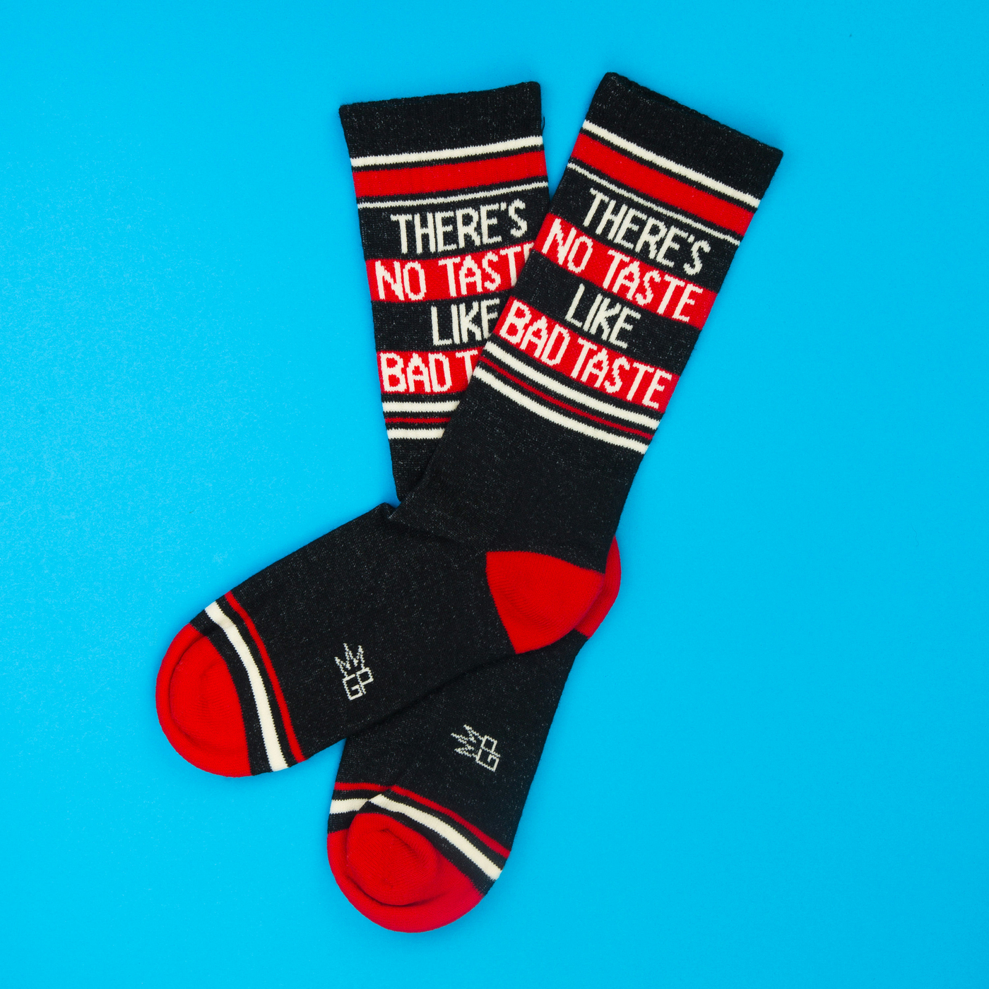 Two dark grey "There's No Taste Like Bad Taste" crew socks with red heels and toes, laid on a blue background.