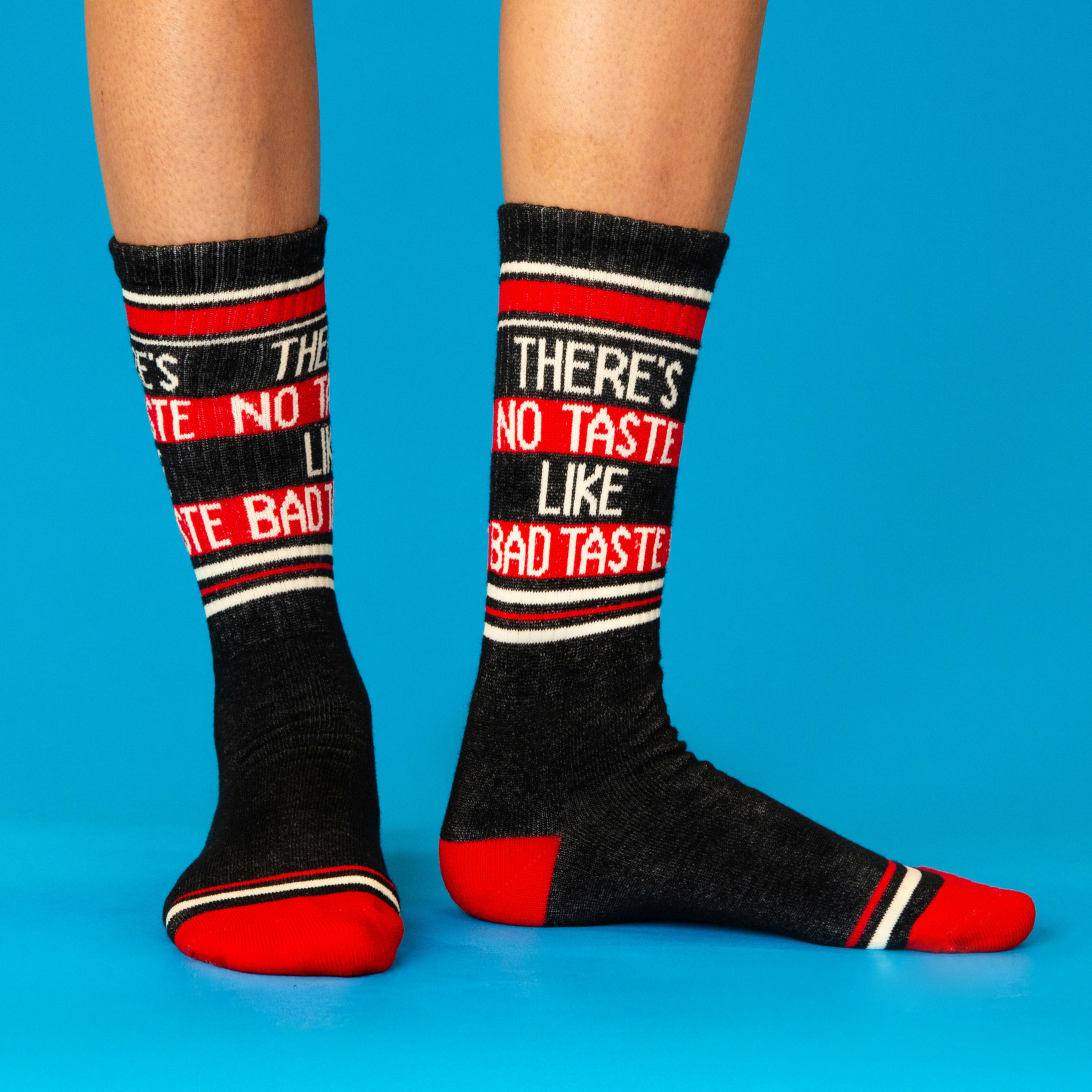 A person's legs and feet wearing dark grey "There's No Taste Like Bad Taste" crew socks on a blue surface.