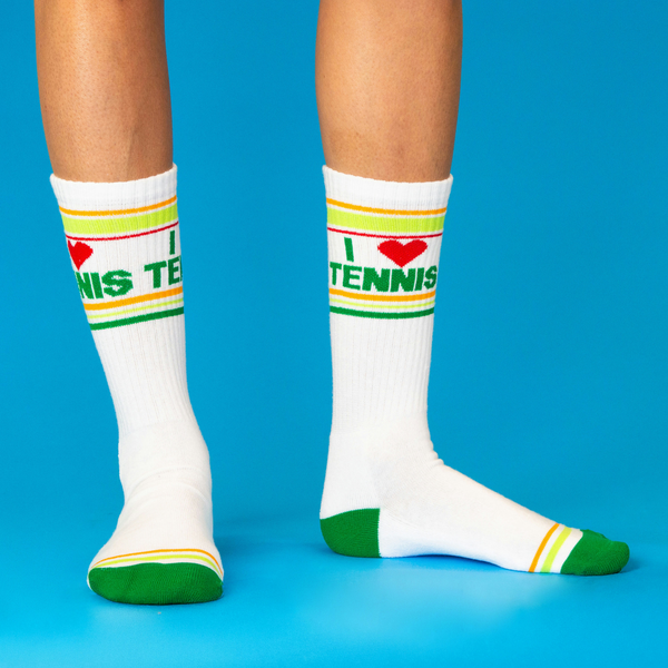 [lf] Two legs wearing white 'I ❤️ Tennis' crew socks with green text and stripes, on a bright blue background.