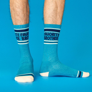 A person's legs wearing teal 'Favorite Brother' crew socks, standing on a bright blue background, viewed from front and side.