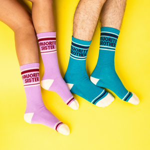 Two pairs of legs on a yellow background, one wearing teal 'Favorite Brother' socks, the other in lavender 'Favorite Sister' socks.