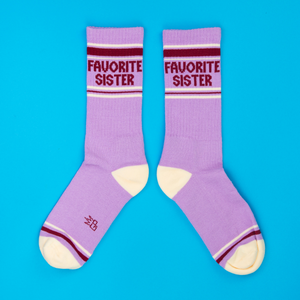 Pair of lavender crew socks with 'Favorite Sister' text, cream heels/toes, and maroon stripes on blue background.