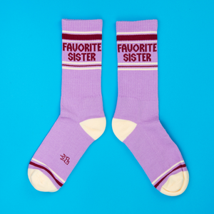 Pair of lavender crew socks with 'Favorite Sister' text, cream heels/toes, and maroon stripes on blue background.