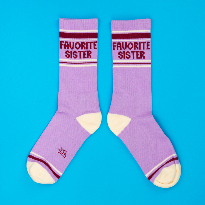 Pair of lavender crew socks with 'Favorite Sister' text, cream heels/toes, and maroon stripes on blue background.