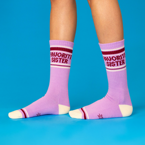 Person wearing lavender 'Favorite Sister' crew socks with cream heels/toes and maroon stripes, standing on blue surface.