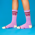 Person wearing lavender 'Favorite Sister' crew socks with cream heels/toes and maroon stripes, standing on blue surface.