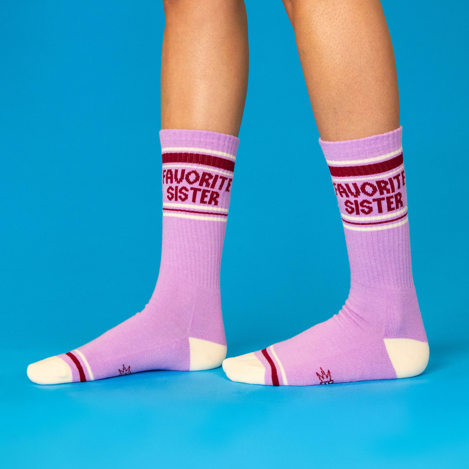 Person wearing lavender 'Favorite Sister' crew socks with cream heels/toes and maroon stripes, standing on blue surface.