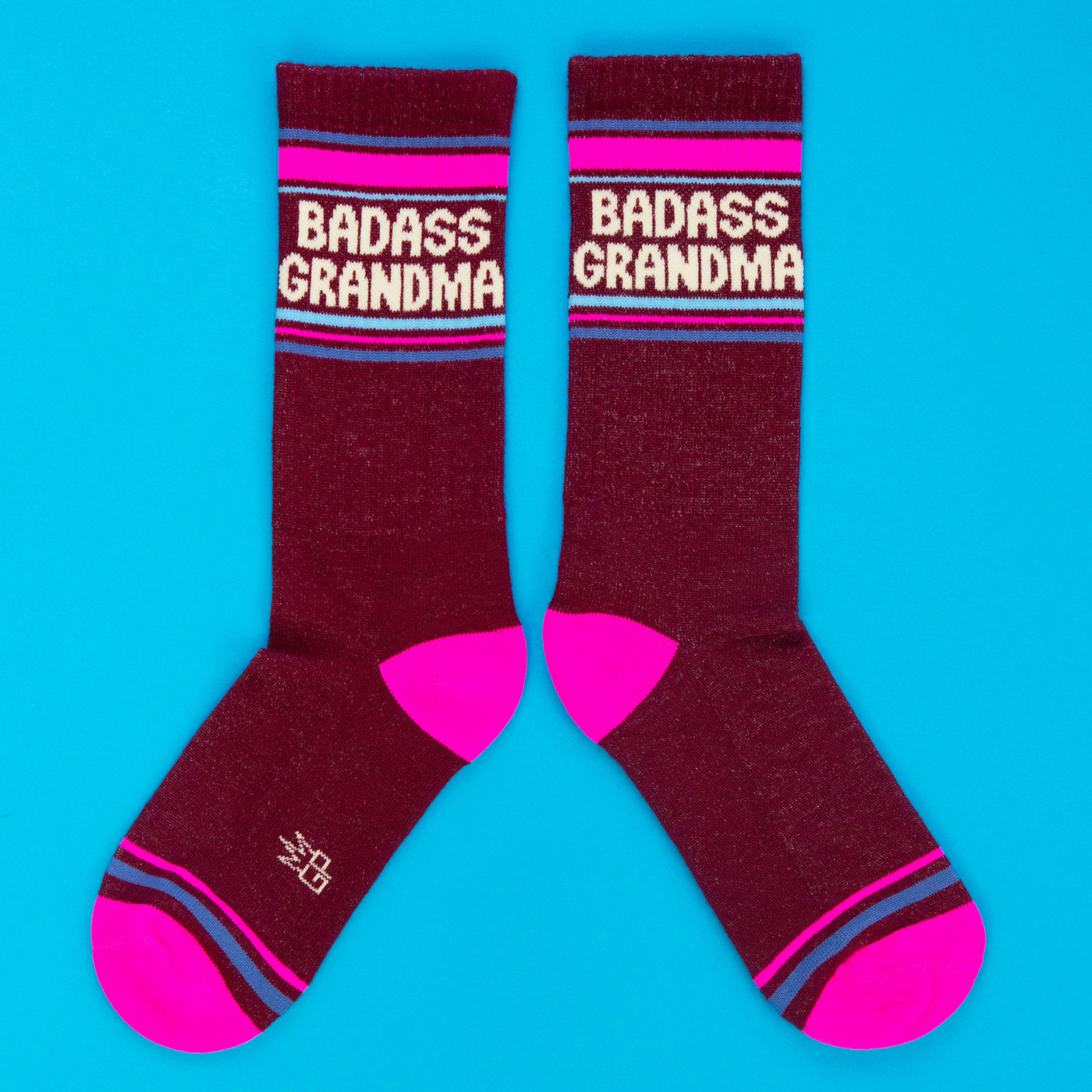 A pair of burgundy "Badass Grandma" crew socks, with hot pink toes and striped cuffs, displayed flat on a bright blue background.