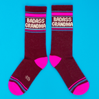 A pair of burgundy "Badass Grandma" crew socks, with hot pink toes and striped cuffs, displayed flat on a bright blue background.