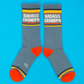Two grey-blue crew socks with "BADASS GRANDPA" text, yellow cuffs, orange heels & toes, and grey logo, on blue background.