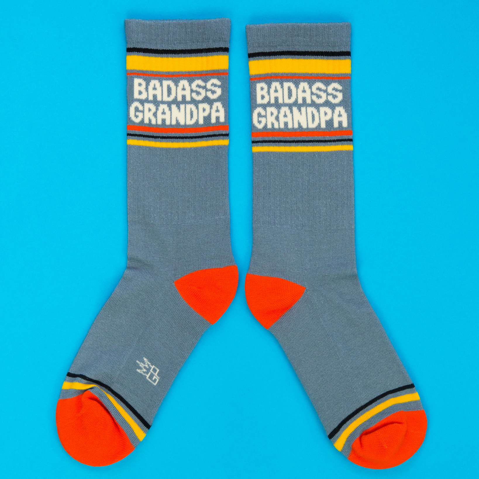 Two grey-blue crew socks with "BADASS GRANDPA" text, yellow cuffs, orange heels & toes, and grey logo, on blue background.
