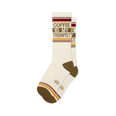 Off-white crew socks reading COFFEE IS MY THERAPIST in brown. Features brown heels, toes, and retro red and yellow stripes.