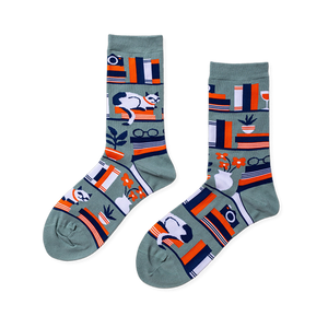 Pair of crew socks in sage green with a bookshelf pattern. Design includes books, a white cat, plants, and reading glasses.