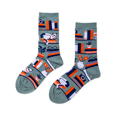 Pair of crew socks in sage green with a bookshelf pattern. Design includes books, a white cat, plants, and reading glasses.