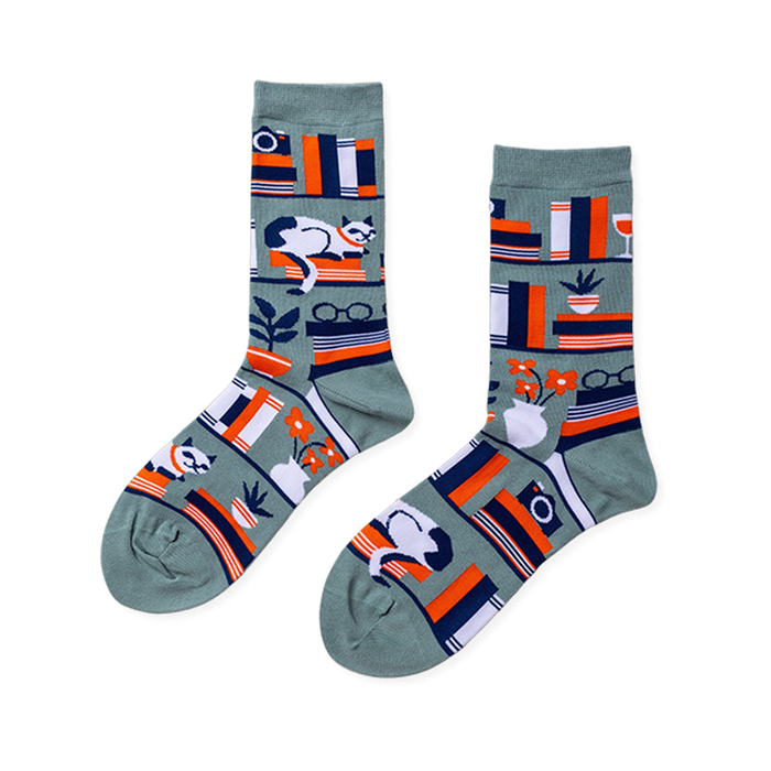 Pair of crew socks in sage green with a bookshelf pattern. Design includes books, a white cat, plants, and reading glasses.