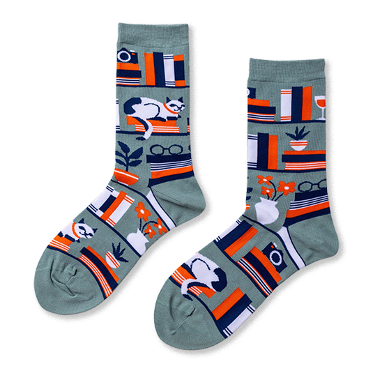 Pair of crew socks in sage green with a bookshelf pattern. Design includes books, a white cat, plants, and reading glasses.