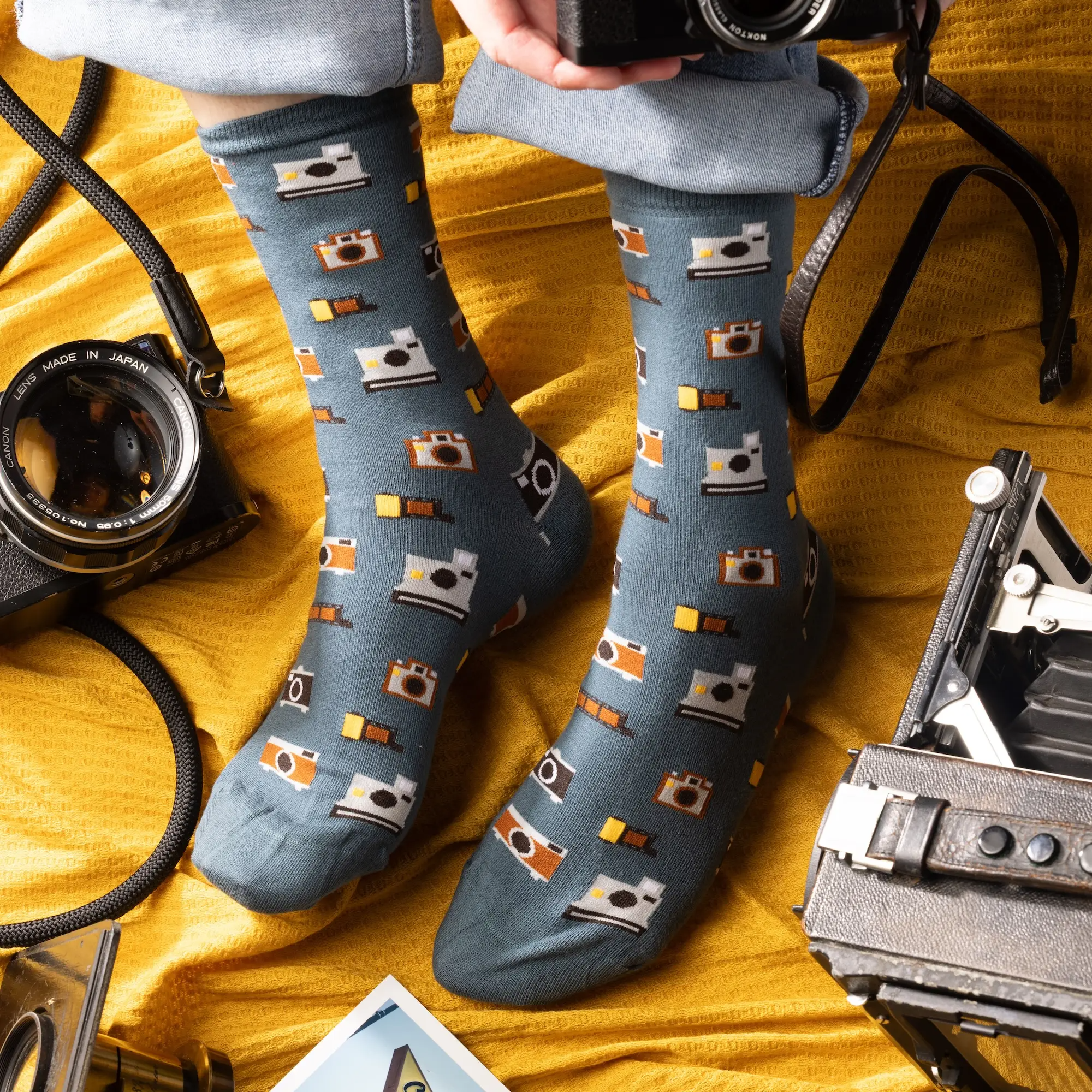 Person wearing blue camera socks and cuffed jeans, sitting on yellow fabric surrounded by real vintage cameras.