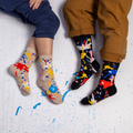Two people wearing black and beige paint splatter socks sitting on a white floor dotted with real wet blue paint.