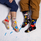 Two people wearing black and beige paint splatter socks sitting on a white floor dotted with real wet blue paint.