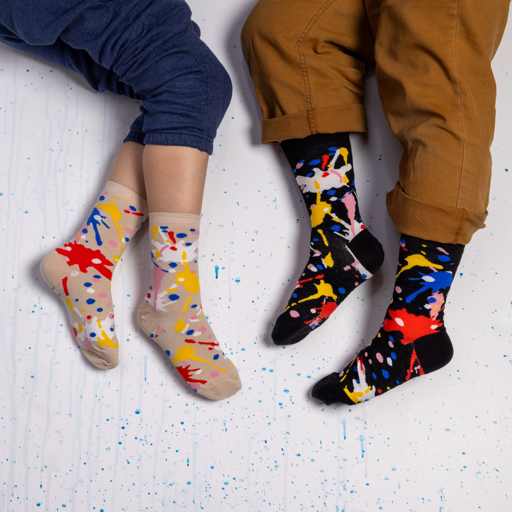 Two people resting on a white floor wearing paint splatter socks. One wears beige socks, the other wears a black version.