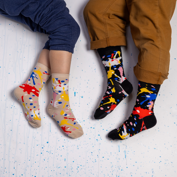 [lf] Two people resting on a white floor wearing paint splatter socks. One wears beige socks, the other wears a black version.