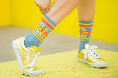 Lifestyle shot: Person wearing 'It's Ok to Fart' socks with gold shoes against a yellow background. [lf]