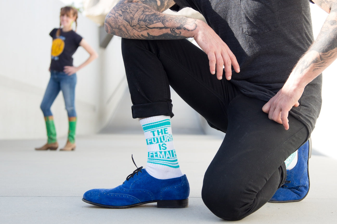 A person kneeling, showcasing the socks and blue shoes; another person is blurred in the background. [lf]