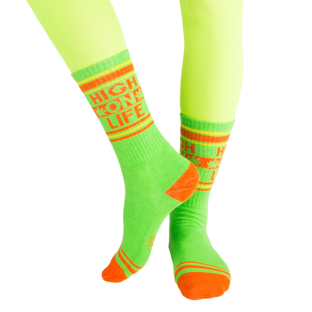Lifestyle shot of 'High On Life' socks on a model's legs; vibrant lime green socks with orange details against a light background. [lf]
