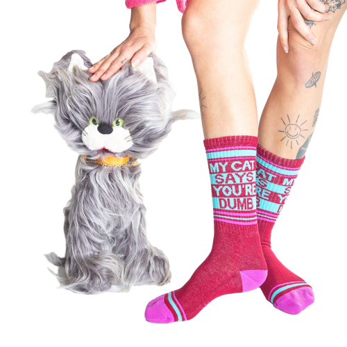 Lifestyle photo showing a model wearing the 'My Cat Says You're Dumb' socks with a fluffy gray cat toy.