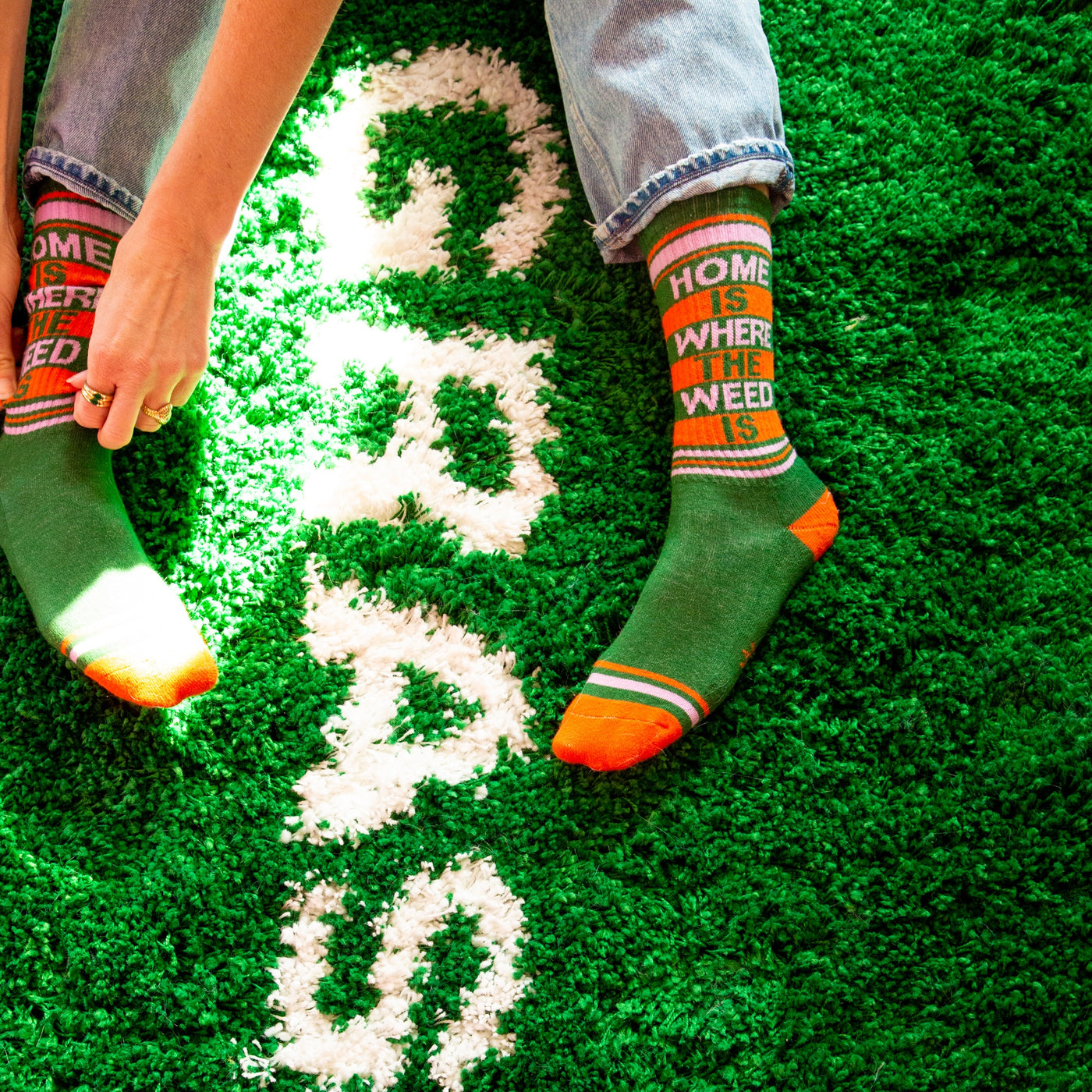 Lifestyle shot of a person wearing the 'Home Is Where The Weed Is' socks, sitting on a green shag carpet. [lf]
